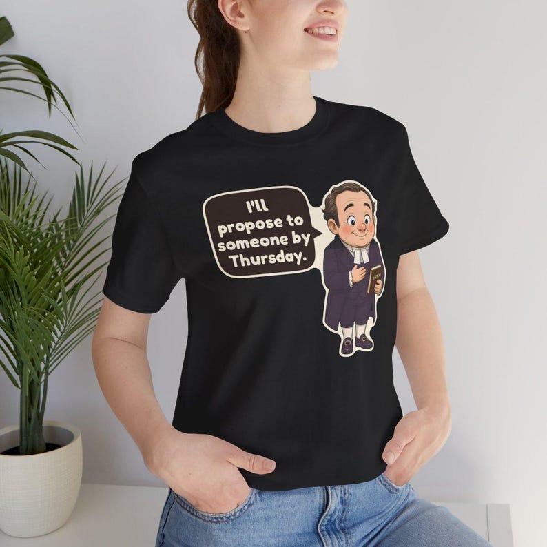 May include: A black short-sleeved t-shirt featuring a cartoon illustration of a man in a dark purple coat and white cravat holding a book. A speech bubble next to the figure contains the text: I'll propose to someone by Thursday.