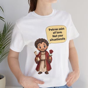 Puede incluir: Una camiseta blanca con una ilustraci&oacute;n de dibujos animados de una figura barbuda con t&uacute;nica roja que sostiene un pergamino y una rosa roja. Un globo de texto amarillo contiene el texto negro: Patron saint of love. Not your situationship.