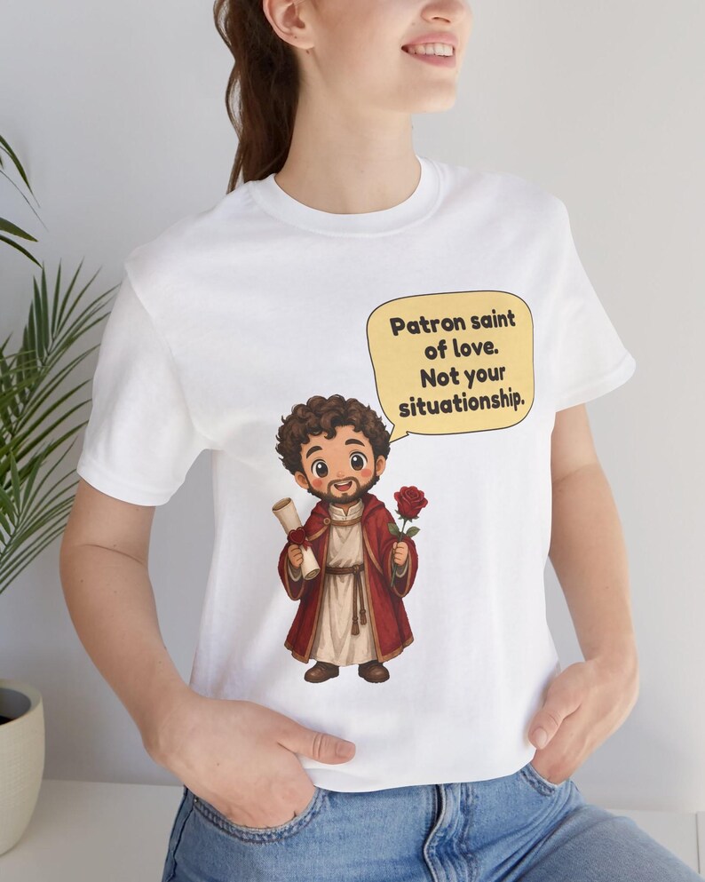 Puede incluir: Una camiseta blanca con una ilustraci&oacute;n de dibujos animados de una figura barbuda con t&uacute;nica roja que sostiene un pergamino y una rosa roja. Un globo de texto amarillo contiene el texto negro: Patron saint of love. Not your situationship.
