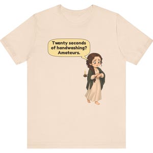 Op de afbeelding: Een lichtbeige t-shirt met een cartoonillustratie van een persoon met lang bruin haar in een vlecht, gekleed in een cr&egrave;mekleurige jurk en donkere sjaal, met een kaars. Een tekstballon bevat de tekst: Twenty seconds of handwashing? Amateurs.