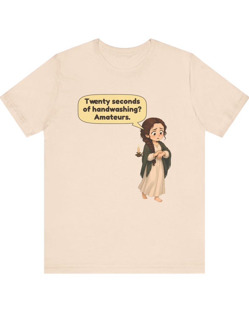 Op de afbeelding: Een lichtbeige t-shirt met een cartoonillustratie van een persoon met lang bruin haar in een vlecht, gekleed in een cr&egrave;mekleurige jurk en donkere sjaal, met een kaars. Een tekstballon bevat de tekst: Twenty seconds of handwashing? Amateurs.