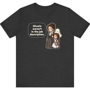 Op de afbeelding: Een donkergrijs t-shirt met een afbeelding van een persoon in een lange donkere jurk met een kaars, een kind met een pop en een klein wit spookje. Een tekstballon bevat de tekst: Ghosts weren't in the job description.