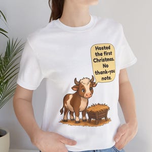 以下が含まれることがあります： 白いTシャツに、干し草が入った木製の飼い葉桶の横に立つ茶色と白の子牛が描かれています。子牛の上の黄色い吹き出しには、黒い文字で「Hosted the first Christmas. No thank-you note.」と書かれています。