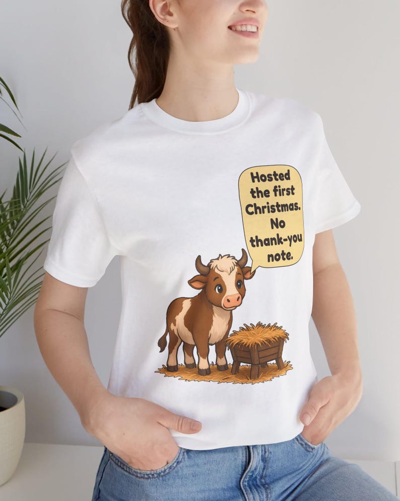 以下が含まれることがあります： 白いTシャツに、干し草が入った木製の飼い葉桶の横に立つ茶色と白の子牛が描かれています。子牛の上の黄色い吹き出しには、黒い文字で「Hosted the first Christmas. No thank-you note.」と書かれています。