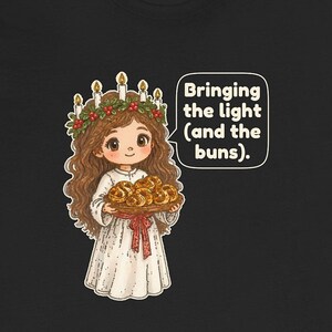 May include: A black t-shirt featuring a cartoon illustration of a figure wearing a white robe and a candle crown with red berries, holding a tray of golden buns. A speech bubble contains the text: Bringing the light (and the buns).