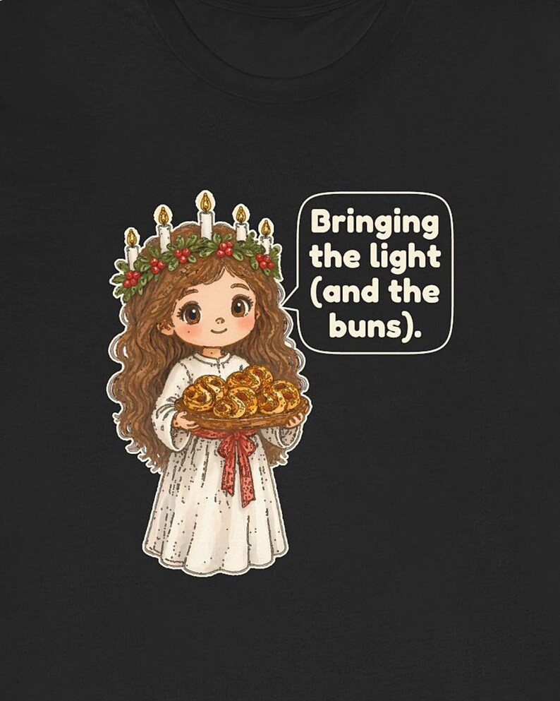May include: A black t-shirt featuring a cartoon illustration of a figure wearing a white robe and a candle crown with red berries, holding a tray of golden buns. A speech bubble contains the text: Bringing the light (and the buns).