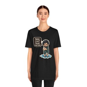 Może przedstawiać: Czarny t-shirt z rysunkową ilustracją brodatej osoby z kręconymi włosami stojącej w kałuży wody. Dymek obok postaci zawiera tekst: Same river? Never met her.
