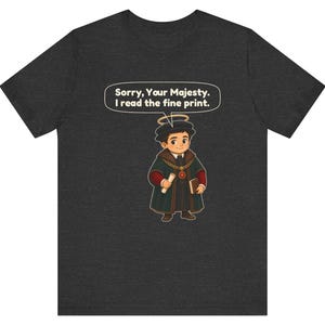 May include: A dark gray t-shirt featuring a cartoon character wearing a dark green robe, a gold chain, and a halo. The character holds a scroll and a book. A speech bubble above the character contains the text: Sorry, Your Majesty. I read the fine print.
