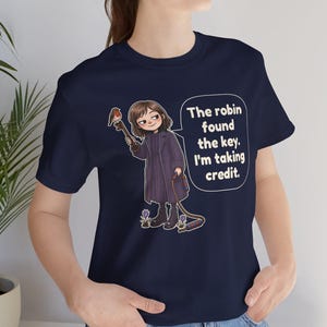 May include: A navy blue t-shirt featuring a cartoon illustration of a person in a dark coat holding a large key with a robin perched on it. A speech bubble contains the text: The robin found the key. I'm taking credit. Small purple flowers are at the feet.