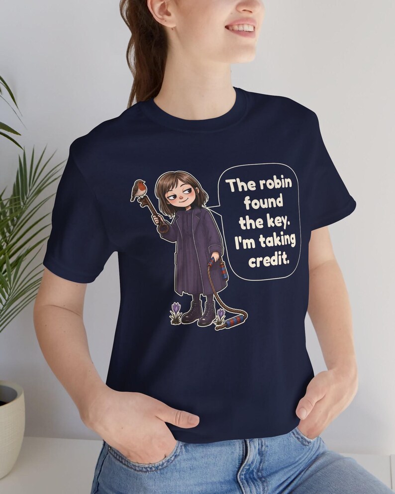 May include: A navy blue t-shirt featuring a cartoon illustration of a person in a dark coat holding a large key with a robin perched on it. A speech bubble contains the text: The robin found the key. I'm taking credit. Small purple flowers are at the feet.