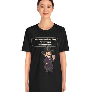 Pu&ograve; includere: Una t-shirt nera con un'illustrazione a cartone animato di un uomo in abito scuro, cappello e baffi che tiene in mano delle carte da gioco. Un fumetto sopra il personaggio contiene il testo: Thirty seconds of fame. Fifty years of interviews.