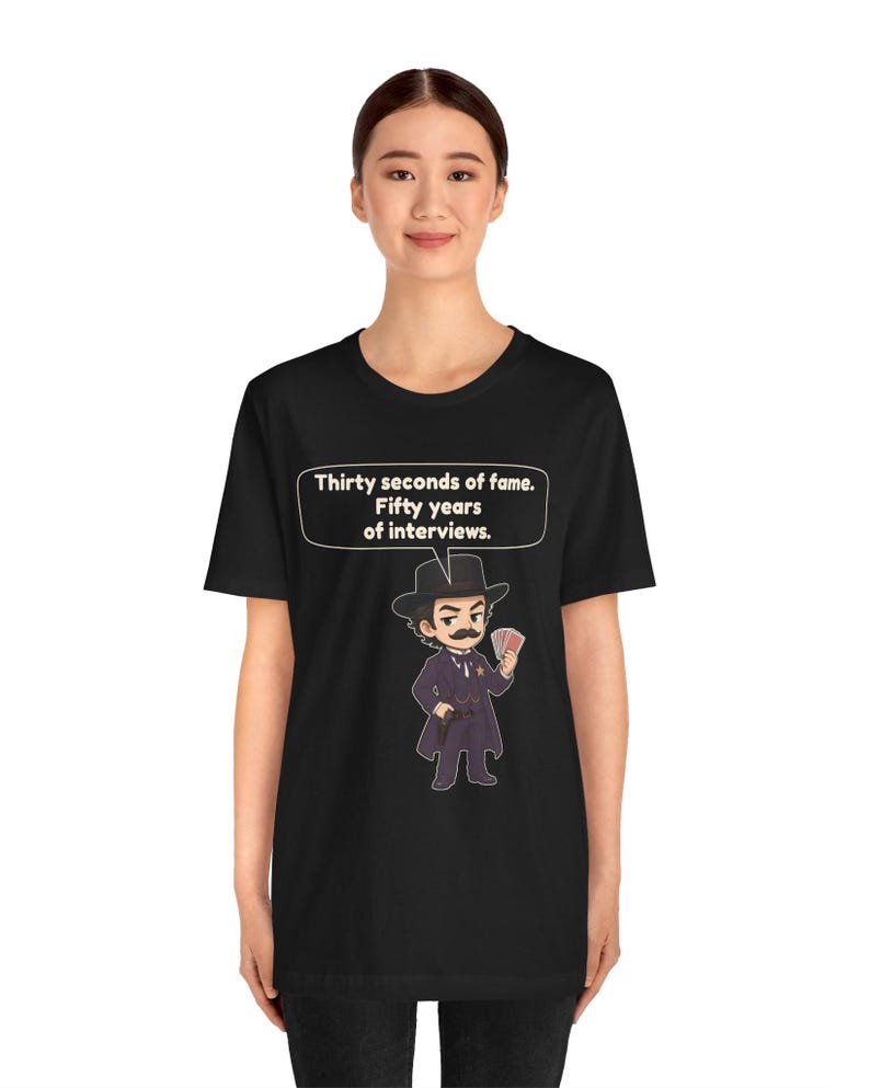 Pu&ograve; includere: Una t-shirt nera con un'illustrazione a cartone animato di un uomo in abito scuro, cappello e baffi che tiene in mano delle carte da gioco. Un fumetto sopra il personaggio contiene il testo: Thirty seconds of fame. Fifty years of interviews.