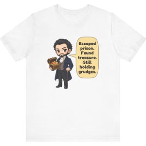 May include: A white t-shirt featuring a cartoon illustration of a bearded character in a dark blue coat holding a treasure chest. A speech bubble next to the character contains the text: Escaped prison. Found treasure. Still holding grudges.