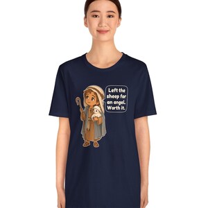 May include: A navy blue t-shirt featuring a cartoon illustration of a shepherd holding a small white sheep. A speech bubble next to the character contains the text: Left the sheep for an angel. Worth it. The design uses brown, blue, and white colors.