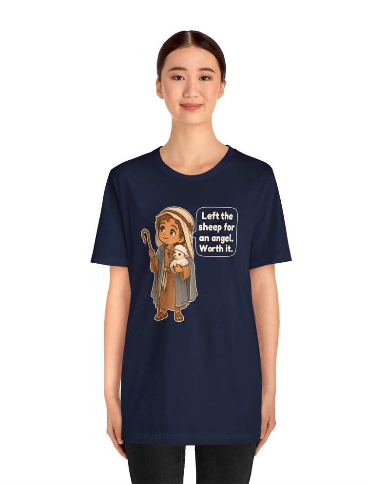 May include: A navy blue t-shirt featuring a cartoon illustration of a shepherd holding a small white sheep. A speech bubble next to the character contains the text: Left the sheep for an angel. Worth it. The design uses brown, blue, and white colors.