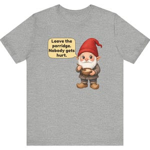 May include: A heather grey t-shirt featuring a graphic of a gnome wearing a red hat and brown clothing, holding a wooden bowl and spoon. A speech bubble next to the gnome contains the text: Leave the porridge. Nobody gets hurt.