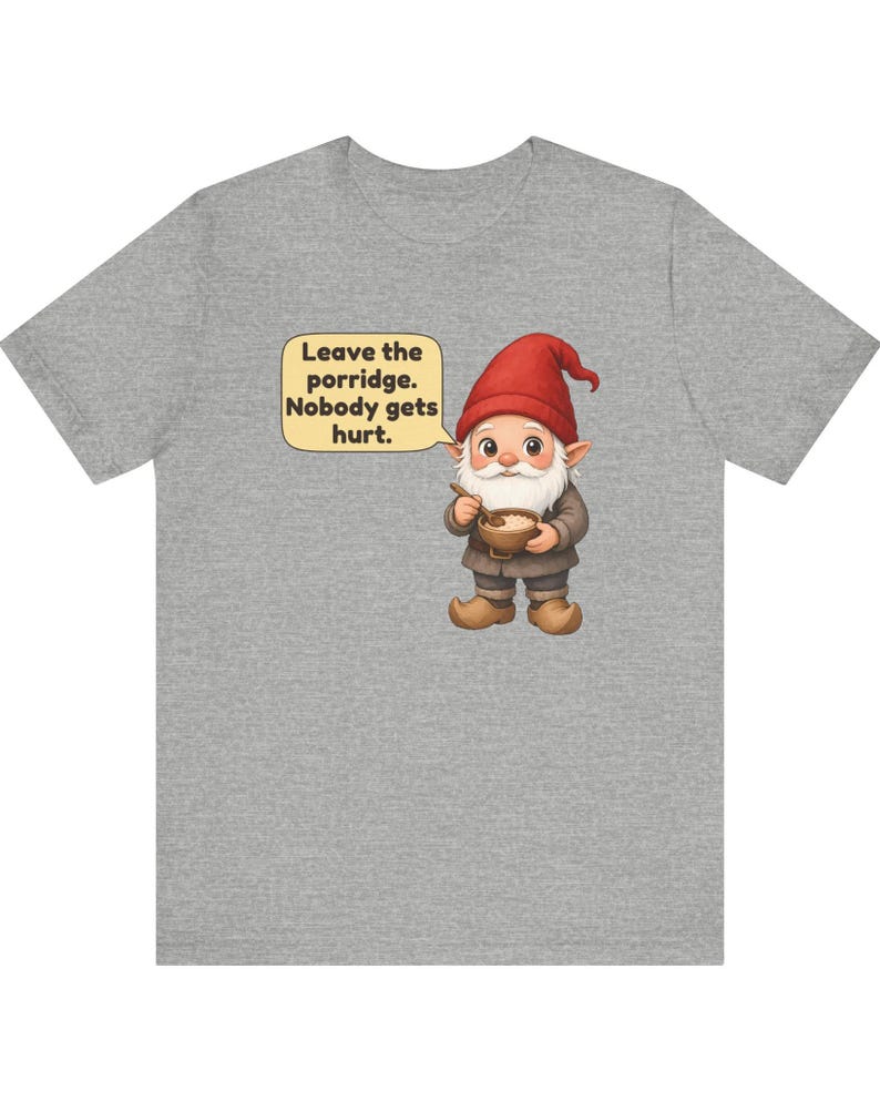 May include: A heather grey t-shirt featuring a graphic of a gnome wearing a red hat and brown clothing, holding a wooden bowl and spoon. A speech bubble next to the gnome contains the text: Leave the porridge. Nobody gets hurt.