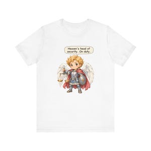 May include: A white t-shirt featuring a graphic of a curly-haired angel in silver armor with white wings, a red cape, a shield, and a golden scale. A speech bubble above the angel contains the text: Heaven's head of security. On duty.