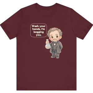 May include: A maroon t-shirt featuring a cartoon illustration of a man in a suit holding a glass flask. A speech bubble above the figure contains the text: Wash your hands. I'm begging you. The design is centered on the front of the garment.