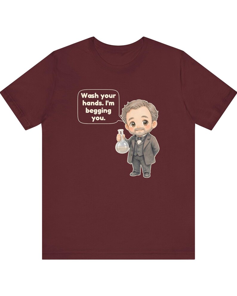 May include: A maroon t-shirt featuring a cartoon illustration of a man in a suit holding a glass flask. A speech bubble above the figure contains the text: Wash your hands. I'm begging you. The design is centered on the front of the garment.