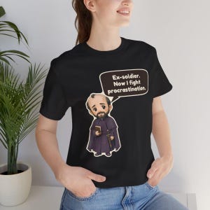 May include: A black short-sleeve t-shirt featuring a cartoon illustration of a bearded person in a dark purple cloak holding a book and a sword. A speech bubble above the character contains the text: Ex-soldier. Now I fight procrastination.