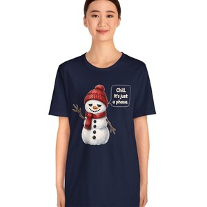 Pode incluir: Uma t-shirt azul-marinho de manga curta com o gr&aacute;fico de um boneco de neve a usar um gorro e um cachecol de malha vermelhos. O boneco de neve tem uma express&atilde;o c&eacute;tica e um bal&atilde;o de fala com o texto branco: Chill. It's just a phase.