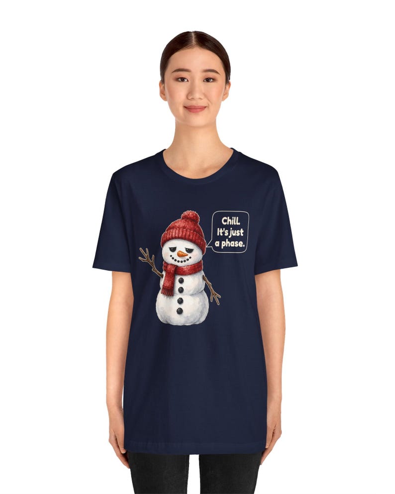Pode incluir: Uma t-shirt azul-marinho de manga curta com o gr&aacute;fico de um boneco de neve a usar um gorro e um cachecol de malha vermelhos. O boneco de neve tem uma express&atilde;o c&eacute;tica e um bal&atilde;o de fala com o texto branco: Chill. It's just a phase.