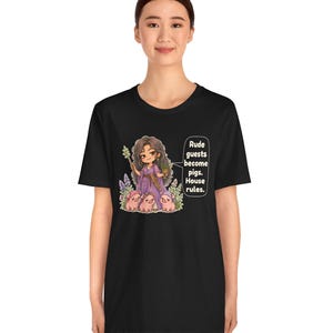 May include: A black t-shirt featuring a graphic of a character in a purple dress holding a wand and a potted plant, surrounded by three small pigs. A speech bubble contains the text: Rude guests become pigs. House rules.