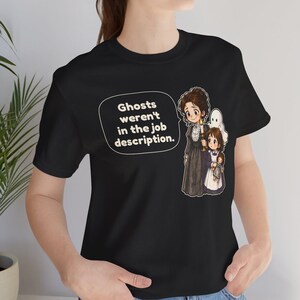 Op de afbeelding: Een zwart t-shirt met een afbeelding van een vrouw en een kind die een kaars vasthouden, vergezeld door een klein wit spookje. Een tekstballon bevat de tekst: Ghosts weren't in the job description. Het ontwerp staat op de voorkant.