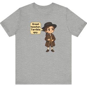 May include: A heather gray t-shirt featuring a cartoon illustration of a person in historical attire wearing a black hat and brown coat, holding a book. A speech bubble next to the character contains the text: Great teacher. Terrible spy.