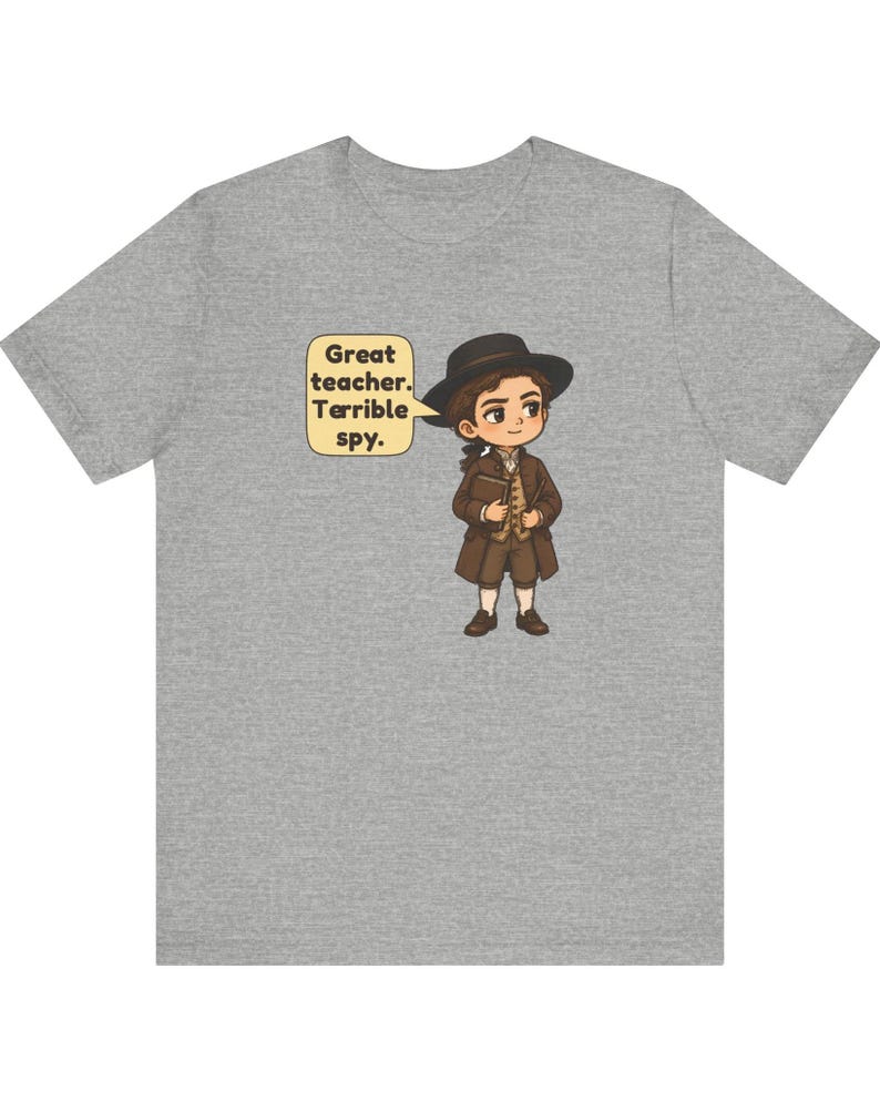 May include: A heather gray t-shirt featuring a cartoon illustration of a person in historical attire wearing a black hat and brown coat, holding a book. A speech bubble next to the character contains the text: Great teacher. Terrible spy.