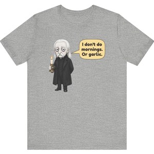May include: A heather gray t-shirt featuring a graphic of a pale, long-haired character in a black coat holding a lit candle. A speech bubble next to the character contains the text: I don't do mornings. Or garlic. The design is centered on the chest.