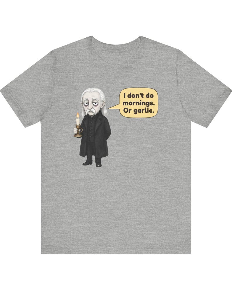 May include: A heather gray t-shirt featuring a graphic of a pale, long-haired character in a black coat holding a lit candle. A speech bubble next to the character contains the text: I don't do mornings. Or garlic. The design is centered on the chest.