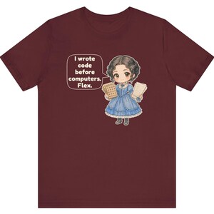 May include: A maroon t-shirt featuring a cartoon illustration of a person in a blue Victorian-style dress holding punch cards and papers. A speech bubble contains the text: I wrote code before computers. Flex.