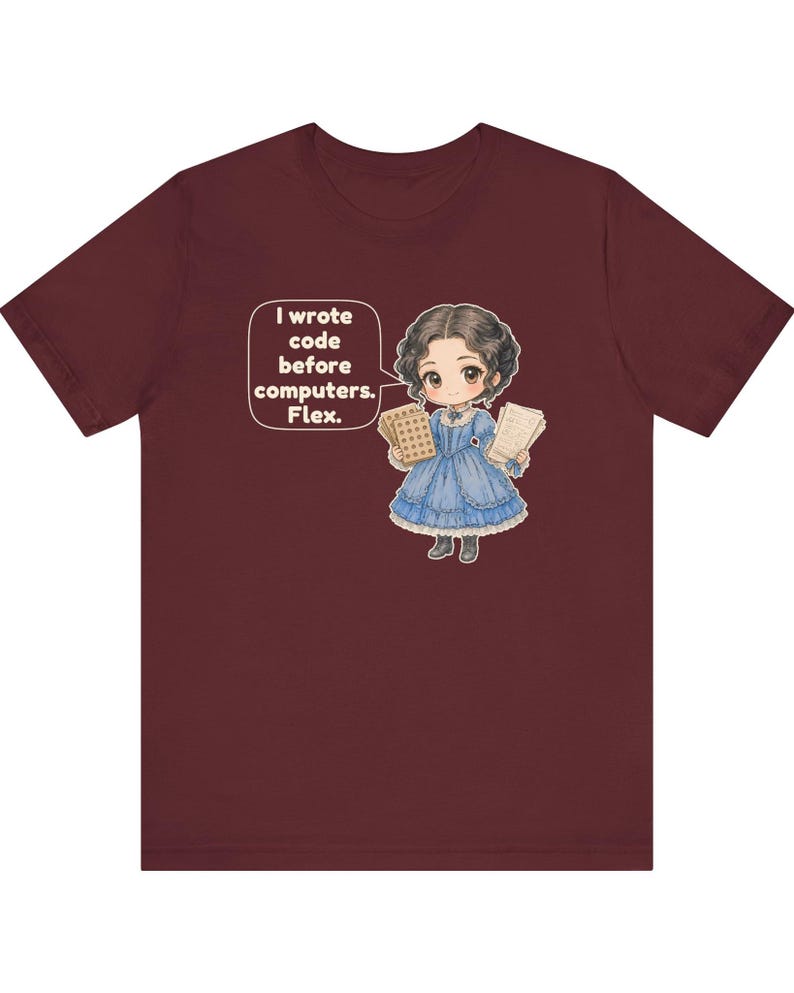 May include: A maroon t-shirt featuring a cartoon illustration of a person in a blue Victorian-style dress holding punch cards and papers. A speech bubble contains the text: I wrote code before computers. Flex.