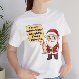 May include: A white short-sleeved t-shirt featuring a cartoon Santa Claus holding a scroll. A yellow speech bubble next to him contains the black text: I know who's been naughty. I keep receipts. The design is centered on the chest area.