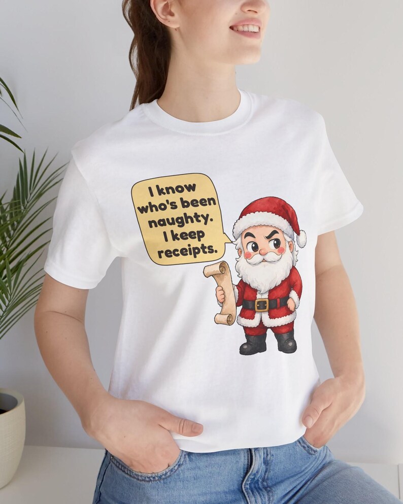 May include: A white short-sleeved t-shirt featuring a cartoon Santa Claus holding a scroll. A yellow speech bubble next to him contains the black text: I know who's been naughty. I keep receipts. The design is centered on the chest area.