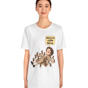 Op de afbeelding: Een wit t-shirt met korte mouwen en een afbeelding van een groot personage dat op de grond ligt, omringd door vele kleine figuurtjes die touwen en ladders gebruiken om het personage vast te binden. Een tekstballon bevat, Sorry, I'm a little tied up.