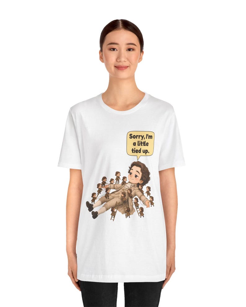 Op de afbeelding: Een wit t-shirt met korte mouwen en een afbeelding van een groot personage dat op de grond ligt, omringd door vele kleine figuurtjes die touwen en ladders gebruiken om het personage vast te binden. Een tekstballon bevat, Sorry, I'm a little tied up.