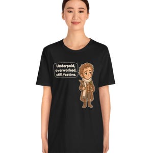 May include: A black t-shirt featuring a cartoon illustration of a person in a brown coat and white scarf holding a quill and candle. A speech bubble next to the character contains the text: Underpaid, overworked, still festive.