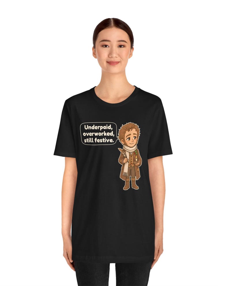 May include: A black t-shirt featuring a cartoon illustration of a person in a brown coat and white scarf holding a quill and candle. A speech bubble next to the character contains the text: Underpaid, overworked, still festive.