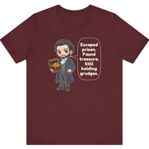 May include: A maroon t-shirt featuring a cartoon illustration of a man in a blue coat holding a treasure chest. A speech bubble next to the character contains the text: Escaped prison. Found treasure. Still holding grudges.