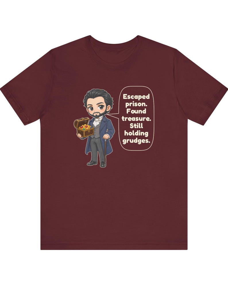 May include: A maroon t-shirt featuring a cartoon illustration of a man in a blue coat holding a treasure chest. A speech bubble next to the character contains the text: Escaped prison. Found treasure. Still holding grudges.