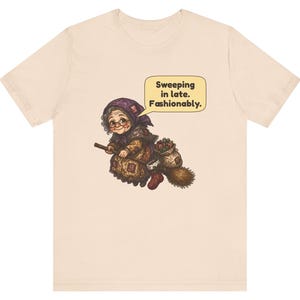 Puede incluir: Una camiseta de color beige claro con un gr&aacute;fico de una persona mayor con un pa&ntilde;uelo morado y ropa estampada, montada en una escoba. Un bocadillo sobre la figura contiene el texto: Sweeping in late. Fashionably.
