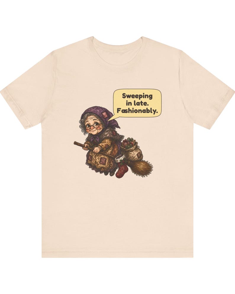 Puede incluir: Una camiseta de color beige claro con un gr&aacute;fico de una persona mayor con un pa&ntilde;uelo morado y ropa estampada, montada en una escoba. Un bocadillo sobre la figura contiene el texto: Sweeping in late. Fashionably.