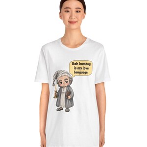 Puede incluir: Una camiseta blanca con el gr&aacute;fico de un personaje de dibujos animados con gorro de dormir y bata gris, sosteniendo una vela encendida y un libro. Un bocadillo contiene el texto: "Bah humbug is my love language."