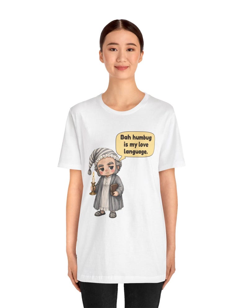 Puede incluir: Una camiseta blanca con el gr&aacute;fico de un personaje de dibujos animados con gorro de dormir y bata gris, sosteniendo una vela encendida y un libro. Un bocadillo contiene el texto: "Bah humbug is my love language."