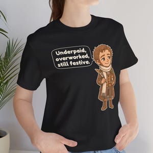 May include: A black t-shirt featuring a cartoon illustration of a character in a brown coat and scarf holding a quill and candle. A speech bubble next to the character contains the text: Underpaid, overworked, still festive.