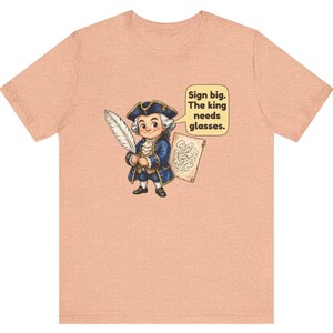 May include: A peach-colored t-shirt featuring a cartoon illustration of a person in 18th-century attire holding a quill and a scroll. A speech bubble contains the text: Sign big. The king needs glasses.