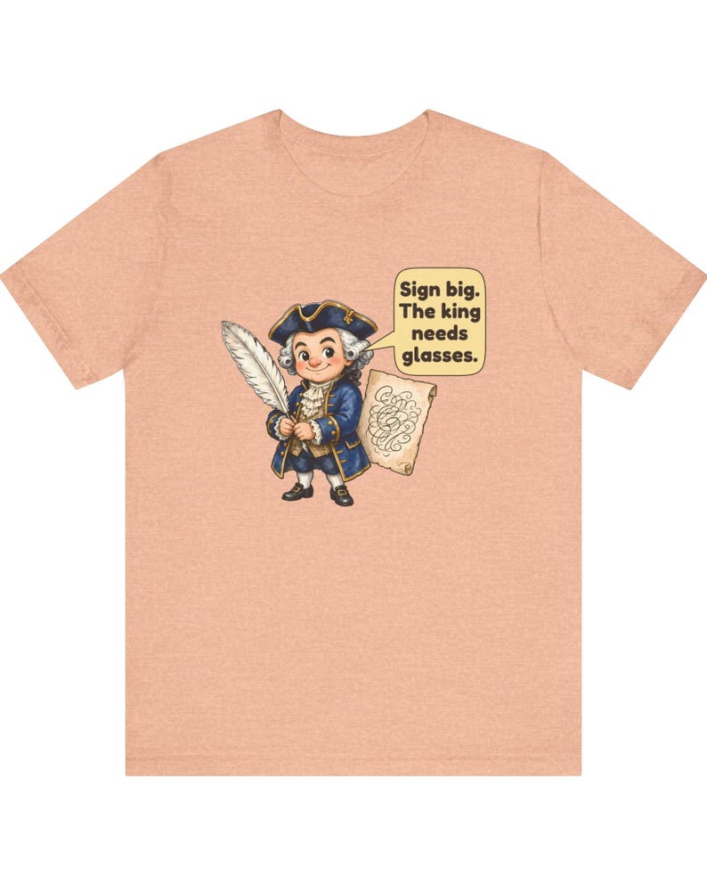 May include: A peach-colored t-shirt featuring a cartoon illustration of a person in 18th-century attire holding a quill and a scroll. A speech bubble contains the text: Sign big. The king needs glasses.
